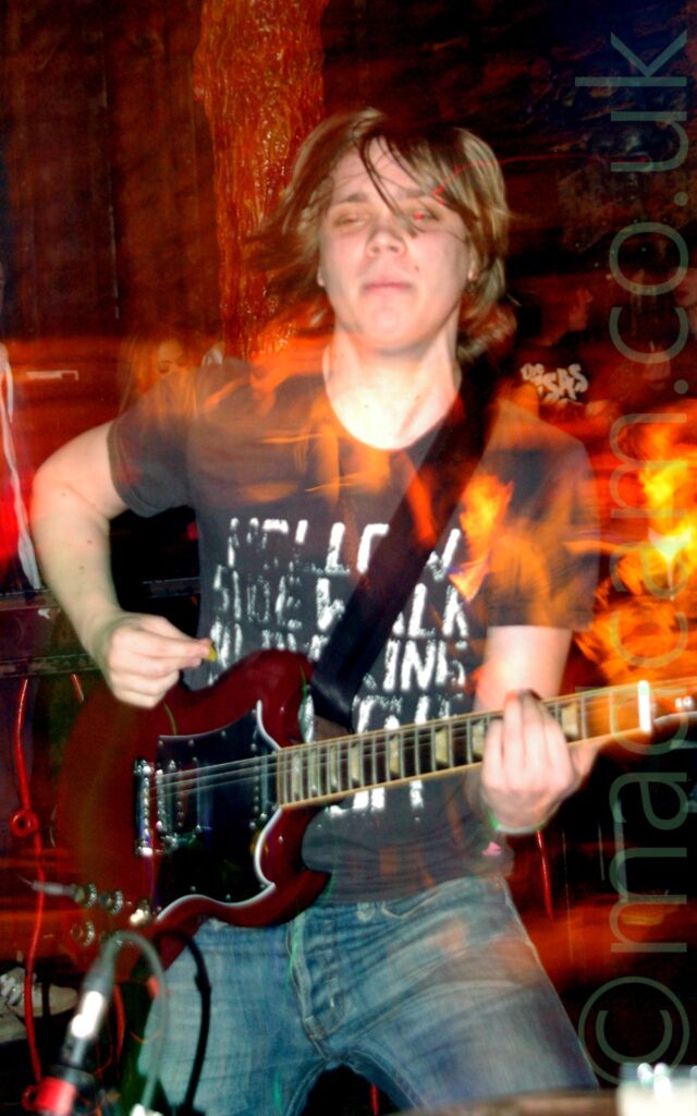 A man with floppy, shoulder-length blond hair, wearing a black t-shirt with faded white text filling the front, and a blue pair of jeans, playing a red electric guitar. An orange pole stretches from floor to ceiling behind him, painted to look sticky and organic and visceral, with some people beyond that, with dark coloured walls behind them. A long exposure and his movement have created red and orange streaks of light and secondary images.