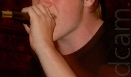Close up of the upper body and face of a young man with short, blond-ish hair, wearing a black t-shirt with a white logo, holding a microphone in his left hand, which he is singing (or possibly snarling) into. He has been afflicted with a terrible case of red eye, caused by the camera flash. There is a red tinge over the entire frame, caused by a long exposure resulting in some movement. Wooden panelling on the wall fills most of the background, with yellow wallpaper showing at the top of the frame.