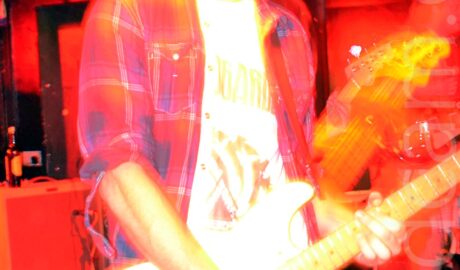 A somewhat over-exposed, long exposure and flashless image of a man with short hair, wearing a red plaid shirt of a white t-shirt and blue jeans, playing a white electronic guitar, with his mouth wide open. Red and yellow stage lights illuminate different parts of the frame, with the walls and ceiling in the background flooded with red light, and his face, arms, and body given a yellow aura. The head of a bass guitar can be seen moving behind his left arm, covered with yellow light.