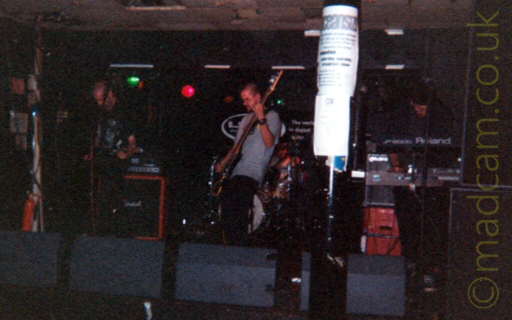 A rather dark and gloomy (and probably over-processed) photo of the 4 members of a band performing on stage. On the left is a man with a shaven head, a black t-shirt with a white logo, and dark trousers, playing an electric guitar. In the middle is a man with short dark hair, wearing a grey t-shirt and black trousers, and playing a bass guitar. Behind him is a man with short dark hair, wearing a white t-shirt, and sitting behind a drum kit. On the right is a man with short dark hair, wearing glasses, and a dark t-shirt and trousers, looking down at the bank of keyboards he is playing. A thick black pole dominates the foreground, slightly off to the right of centre, with some sheets of white paper taped to it, with only the word "Oceansize" discernible at the top. A row of grey monitor speakers line the front of the stage. Black walls and a grey ceiling fill the rest of the frame.