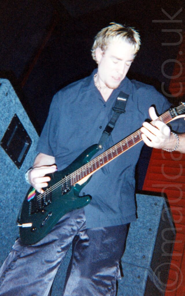 A man with short light-coloured hair, wearing a black shirt and shiny black trousers, and playing a very dark green electric guitar. Stacks of grey cloth-covered speakers stand behind him with black and red walls filling the background.