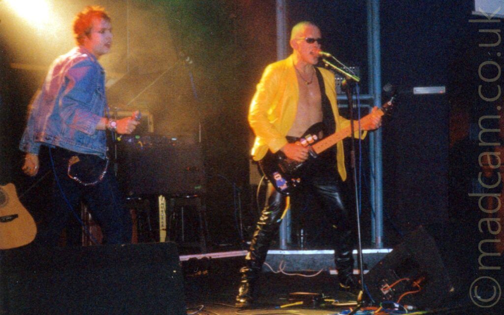 Two men on stage as part of a rock band. On the left is a man with spiky blond hair, wearing a denim jacket and dark blue jeans, holding a microphone in his right hand. On e right is a man with a shaved head, wearing an open yellow pvc jacket that exposes his bare chest, and black pvc trousers, and playing a black electric guitar, while singing into a microphone on a stand. Black wedge monitor speakers are at the front of the stage on both sides of the frame, surrounded by multi-coloured cables. More speakers and various amplifiers are visible at the rear and side of the stage, with black walls in the background, and a yellow beam of light coming from the top left corner.
