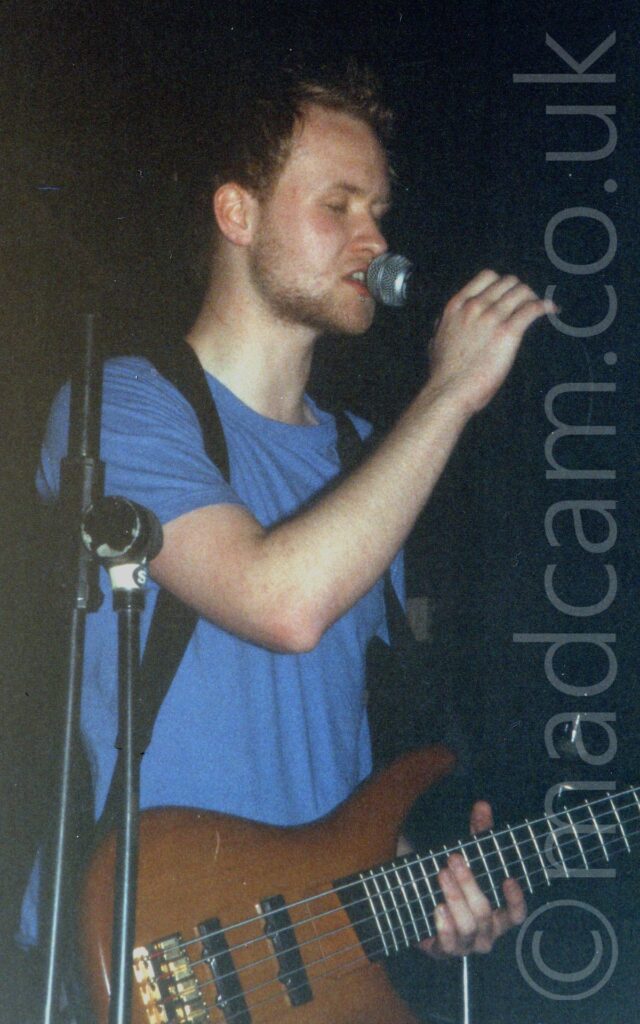 A man with short dark hair and short beard, wearing a blue t-shirt, and with a bass guitar attached to a black strap slung over both shoulders. He is holding on to a microphone on a stand with his right hand, as he sings in to it. Black walls and ceiling fill the rest of the frame.