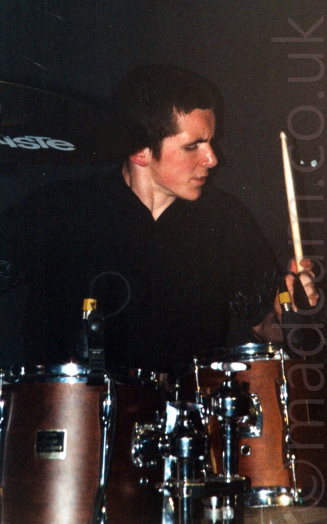 A man with short dark hair, wearing a black shirt, sitting behind a brass-coloured drumkit with a drumstick raised in his left hand, and his head turned to his left. Black walls fill the background.