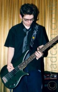 A man with short dark hair and with eyeglasses on his face, wearing a black shirt short sleeved shirt with a knotted grey tie hanging around the shirts open neck, and a dark blue pair of jeans. He is playing a metallic dark green bass guitar, attached to a black and grey strap hanging over his left shoulder. A speaker sits in the background in the bottom right corner. Gold velour curtains cover the wall in the background.