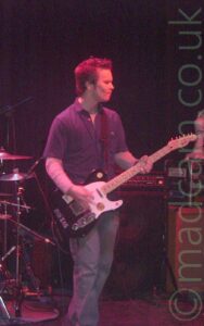 A man performing on a stage as part of a rock band. He has short, slightly spiky dark hair, and is wearing a dark short-sleeved shirt over a long-sleeved, light-coloured t-shirt, as well as a blue pair of jeans. He is playing a black and white electric guitar, while looking off to the right of the frame. Some of the metal supports for some of the drum kit can be seen behind him on the left, while several amplifiers are sitting on the stage behind him on the right. The stage lights are giving the entire frame a red cast, while black curtains right at the back fill the rest of the frame.
