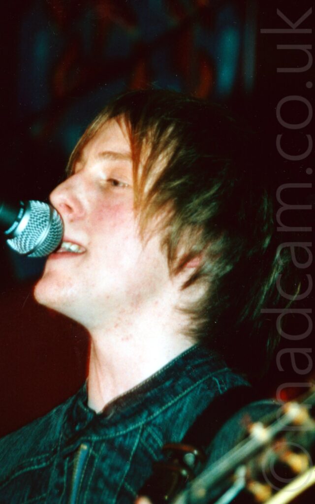 A man with ear-length blond hair, wearing a dark blue denim jacket, singing into a microphone on a stand that is maybe a little bit too tall for him. Black walls fill the background.