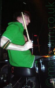 Side view of a man with shaggy dark hair, wearing a green polo shirt, with a green, black, and yellow wristband just below his elbow, sitting behind a black drum kit, his right arm held down by his side but the drumstick raised in the air. His mouth is open as if he is screaming. Black walls fill the background.