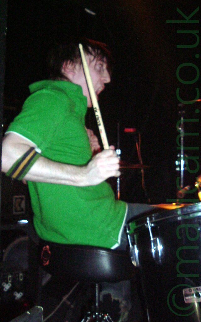 Side view of a man with shaggy dark hair, wearing a green polo shirt, with a green, black, and yellow wristband just below his elbow, sitting behind a black drum kit, his right arm held down by his side but the drumstick raised in the air. His mouth is open as if he is screaming. Black walls fill the background.