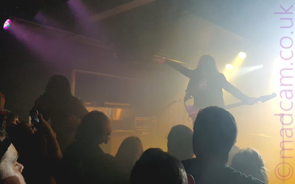 Two men on a stage, performing as part of a band. On the left is a man, silhouetted against the brighter background, hunched over while singing into a microphone in his right hand, leaning out over the audience. On the right is a man, also mostly a silhouette, wearing a denim jacket over a black t-shirt, playing a purple bass guitar, and pointing off into the crowd with his right hand. The heads of people in the first few rows of the pit are visible at the bottom of the frame, while blooms of yellow and white light fill the stage, particularly on the right of the frame.