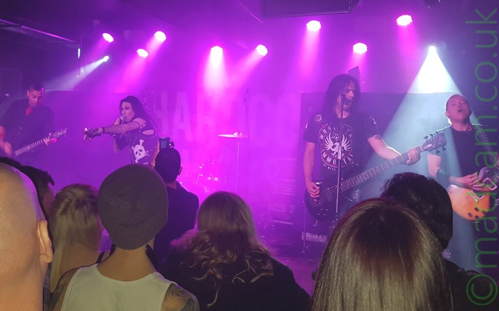 Four members of a band performing on a stage. On the left is a man with short dark hair, wearing a black shirt, and playing a black electric guitar. Next is a woman with long dark hair, holding a microphone to her mouth with her left hand as she sings, while pointing with her left hand at the audience. he has spiked leather gloves on her hands, torn fishnets on her arms, and a black leather jacket with a white skull and crossbones on the front. There are 2 men on the right of the frame, the first has long dark hair, wearing a black t-shirt with a bird with outstretched wings in the middle, surrounded by indistinct text arranged in a circle, playing a 5 string electric bass guitar. To his left, on the right of the frame, is a much smaller man with very short dark hair, wearing a black t-shirt, and playing a varnished wood electric guitar, with his eyes closed as he plays. The backs of the heads of people in the crowd fill the foreground, while almost the entire stage is bathed in purple light from the many purple stage lights. On the right of the frame, a single stage light highlights the guitarist on that side in white light.