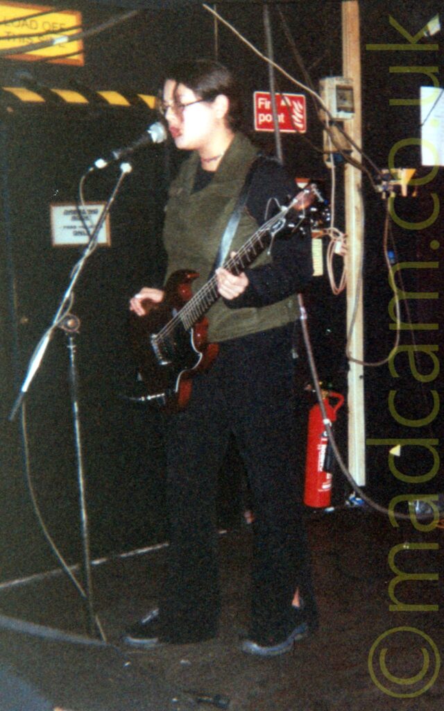 A woman on a stage, playing a black and red electric guitar, and singing into a microphone on a stand in front of her. She has long dark hair that has been tied back neatly behind her ears, with a single clump hanging free just above her wire frame glasses. She is wearing a greeny-grey vest top over a black top and black trousers. Black walls fill the background, with a strip of yellow and black hazard tape on the wall directly behind her.