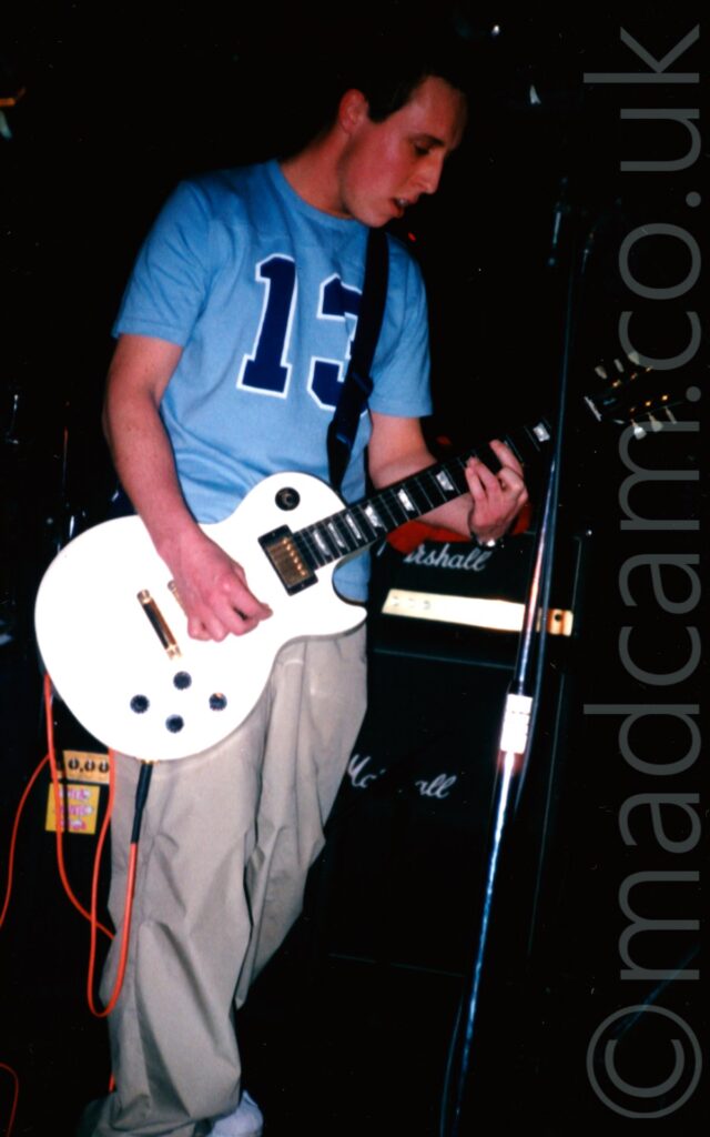 A man with short dark hair, wearing a light blue t-shirt with the large number "13" in the middle in dark blue and outlined in white, and baggy grey jeans, playing a white electric guitar. A microphone stand can be see in the foreground on the right, with stacks of amplifiers in the background, in front of black walls.