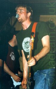 A man with short dark hair and stubbly beard, wearing a green t-shirt with a circular white logo in the middle of the chest, and a blue pair of jeans. He is playing a black bass guitar attached to a bright orange strap over his left shoulder, and leaning in to a microphone on a stand as he sings. Another member of the band can be seen playing a guitar in the bottom left of the background, with gold cloth hangings covering the walls in the rest of the background.