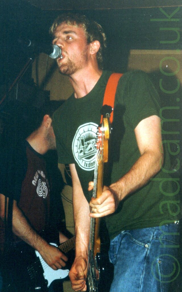 A man with short dark hair and stubbly beard, wearing a green t-shirt with a circular white logo in the middle of the chest, and a blue pair of jeans. He is playing a black bass guitar attached to a bright orange strap over his left shoulder, and leaning in to a microphone on a stand as he sings. Another member of the band can be seen playing a guitar in the bottom left of the background, with gold cloth hangings covering the walls in the rest of the background.