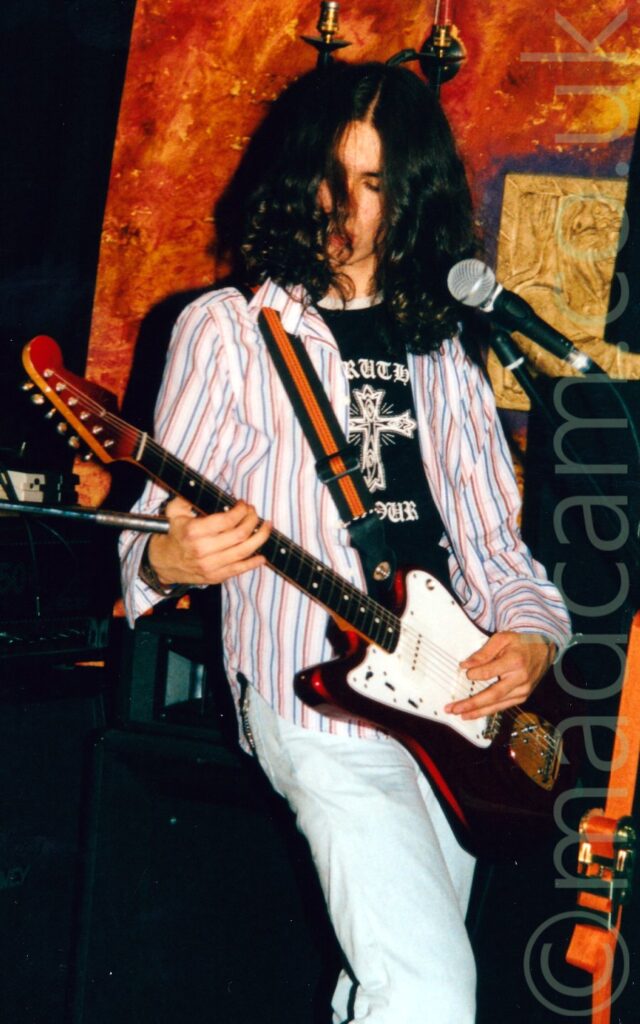 A man with long, wavy, dark hair, wearing a white long-sleeved shirt with red and blue vertical stripes over a black t-shirt with a white Celtic cross on the front. He is playing a black electric guitar left-handed, with a microphone on a stand in front of him. Black walls fill the background, with an orange-painted board behind him.