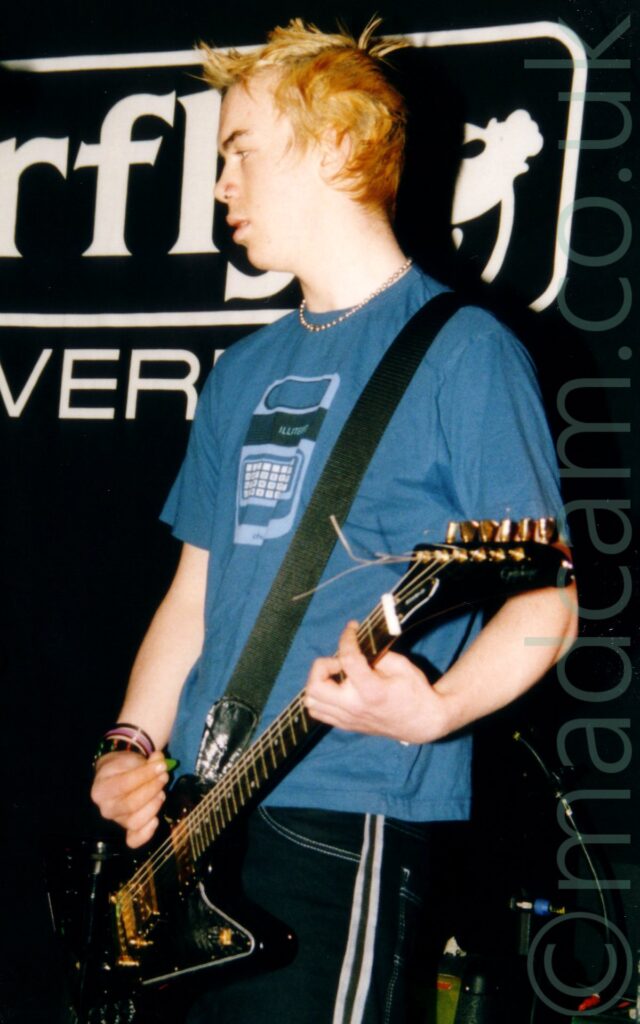 A man with short, blond, spiky hair, wearing a blue t-shirt with a pale blue image of a Texas Instruments Speak 'n Spell on the front, and black jeans with a pair of thin white stripes running down the side of the leg, and playing a black electric guitar, while staring off to the left of the frame. Black walls fill the background, with a white logo of (part of) the word "Barfly" in the white outline of an oblong, over part of the word "Liverpool" behind him on the wall.