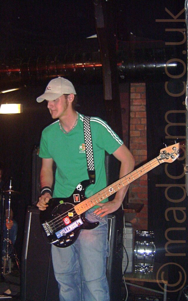 A man wearing a grey baseball cap over his red hair, wearing a green t-shirt and blue jeans, with a black bass guitar hanging from a black and white strap slung over his left shoulder. His left hand is resting on his hip, his right hand on the top of the bass, and he is staring into the distance. Speakers and parts of a drum kit can be seen behind him, with brown brick walls, some covered by black drapes, filling the background.