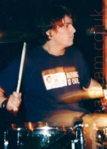A man with ear-length dark hair, wearing a dark blue t-shirt with a white block on the front containing a drawing of a couple of thigures, next to the white text "Nothing is cool." He is sitting behind a black drumkit, a drumstick in his right hand held aloft, while the stick in his left hand is just a blur of motion. A microphone on a stand is pointing at his face on the right of the frame, and he looks as if he is staring at it. Black walls fill the background.