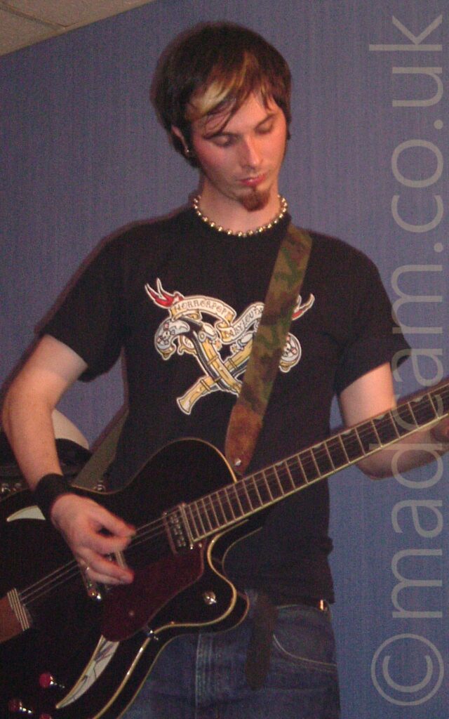 A man with short dark hair, wearing a black t-shirt with a white and yellow logo plastered across the chest, and playing a black electric guitar, performing on a stage. A guitar strap with a green and brown camouflage scheme is hanging over his left shoulder, supporting the guitar. He is looking down at the fingers on his left hand. Purple walls fill the background.