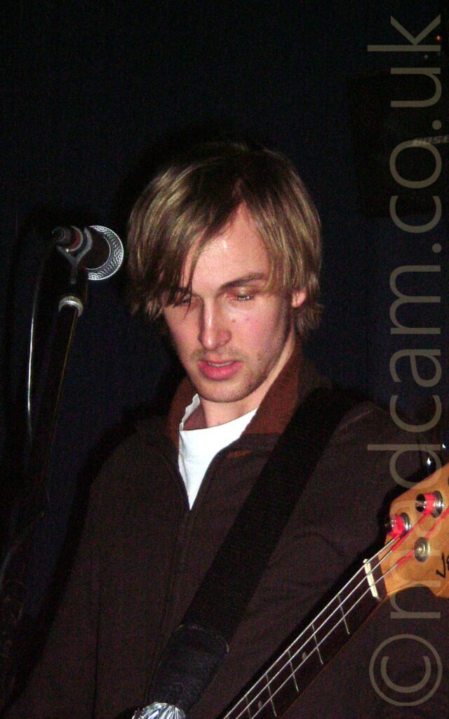 Close-up of the head and shoulders of a man with floppy brown hair, wearing a red light jacket over a white t-shirt, and playing a bass guitar. There is a microphone on a stand in front of his face. Black walls fill the rest of the frame.