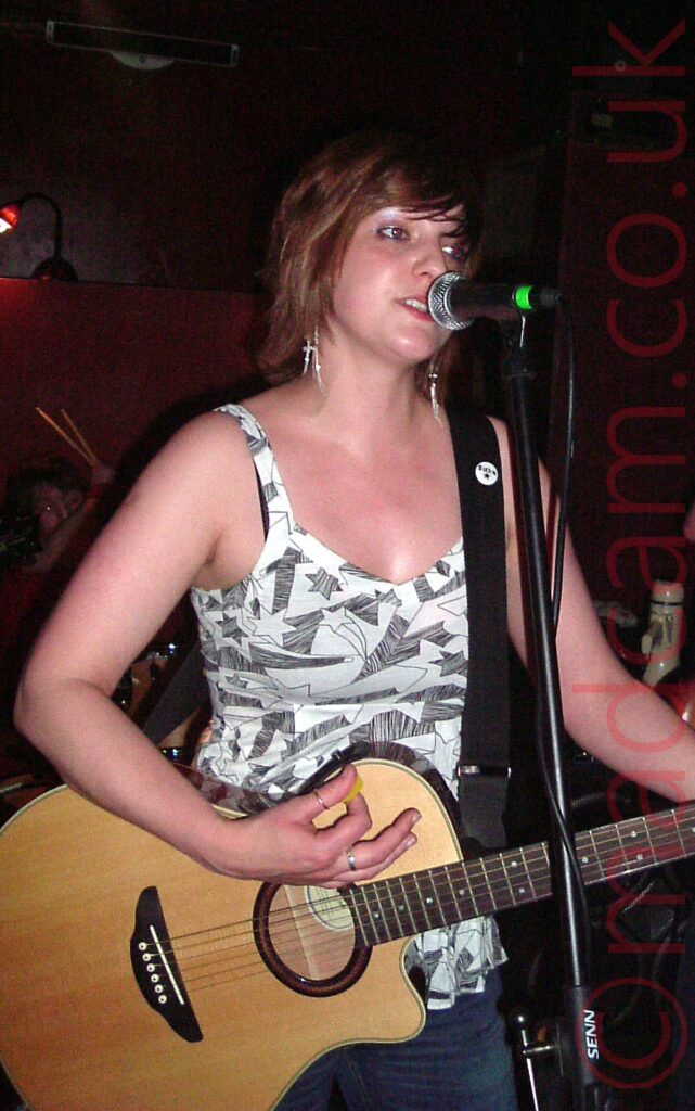 A woman with red hair, wearing a white tank top with a black and grey geometric print, and playing a varnished wood acoustic guitar attached to a black strap over her shoulder, as she sings into a microphone on a stand in front of her. Black walls fill the background.