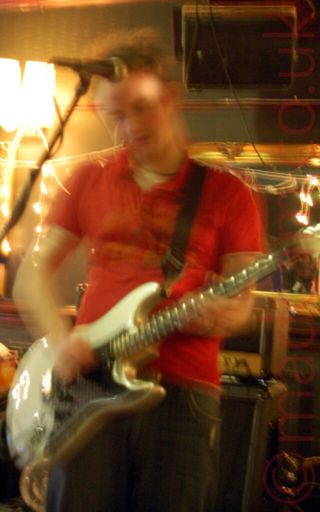 A man with short dark hair, wearing a red polo shirt over a white t-shirt, and blue jeans, and playing a white electric guitar attached to a black strap hanging over his left shoulder. A microphone on a stand hangs down in front of him. A stack of black amplifiers is in the background behind him, with mirrors festooned with lights hanging on the green walls behind him.