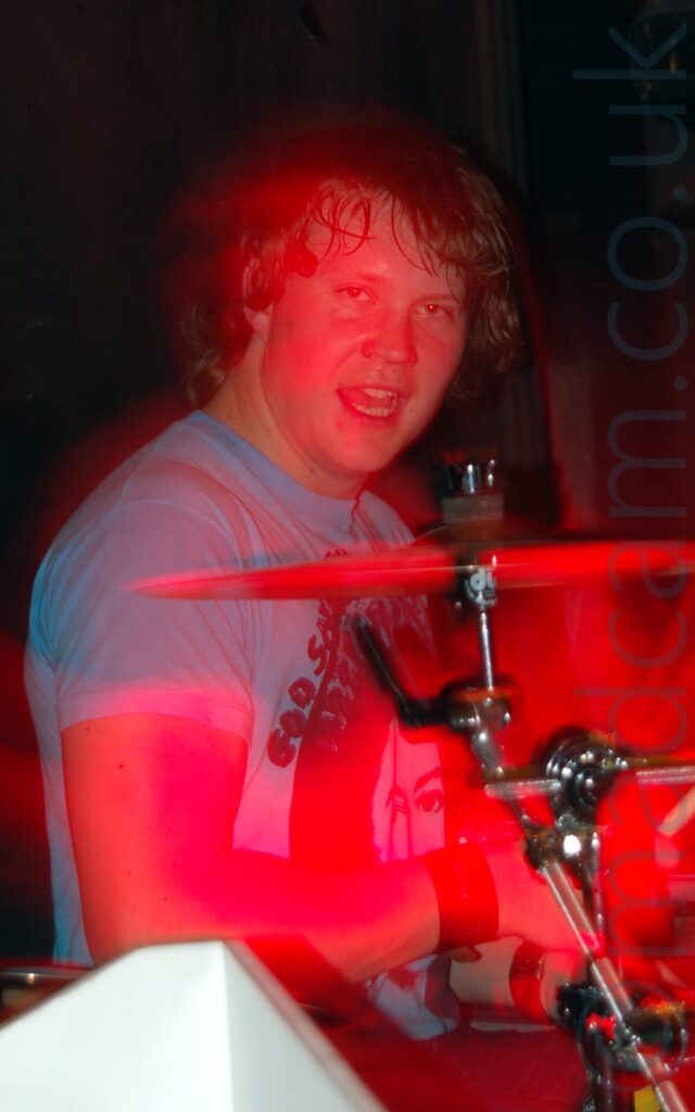A man with shaggy brown hair, wearing a light blue t-shirt with an indistinct black logo on the front, sitting behind a drum kit and staring down the camera lens. Blooms of red light surround his face and the drum kit. Black walls fill the background.