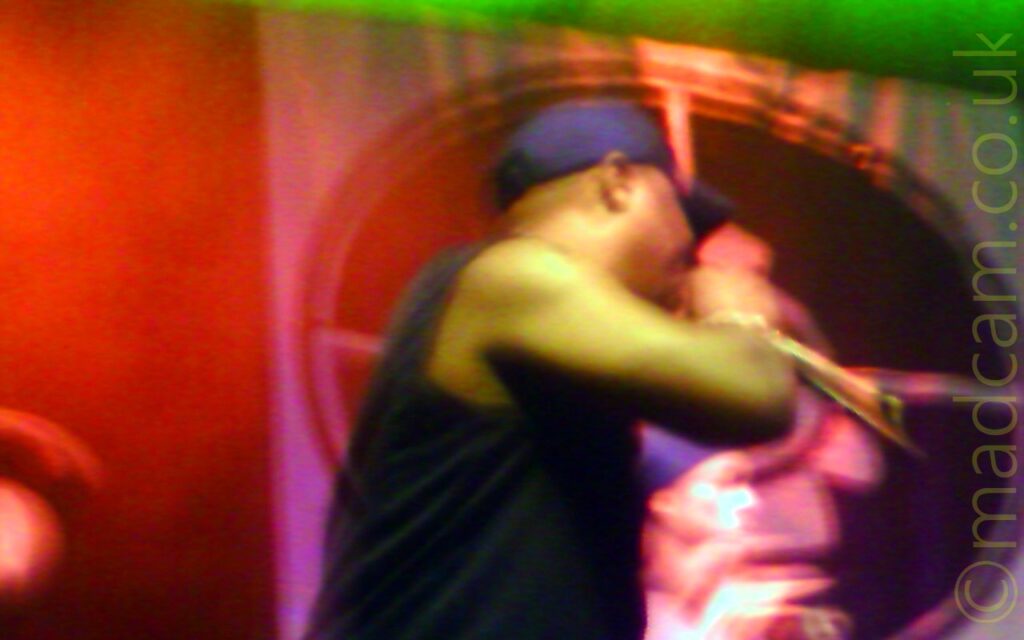 A man wearing a blue baseball cap and a black vest top, holding a microphone in his right hand while turning to his left, away from the camera. A large poster with an image of a figure in a crosshair is on the wall in the background, and has green and red lights shining on it.