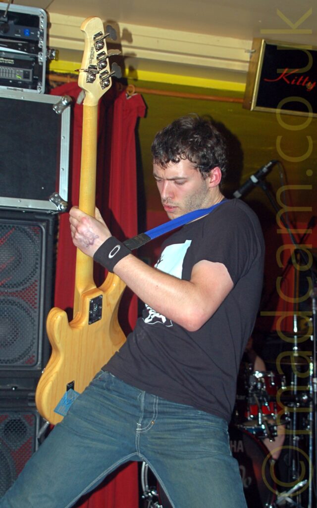 A man with short, slightly wavy dark hair, wearing a black t-shirt and blue jeans, and playing a varnished wood bass guitar, held slightly aloft and off to his right. A floor to ceiling stack of amplifiers is on the left of the background, with a drumkit on the right, and an open red curtain revealing another room behind. Yellow walls and ceiling complete the image.