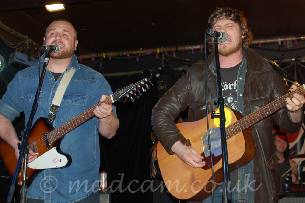 Two men performing on a stage. On the left is a man with close-cropped hair and beard, wearing a blue denim shirt over a black t-shirt, and playing a brown and white electric guitar, and singing into a microphone on a stand in front of him. On the right is a man with long-ish, brown, curly hair, wearing a brown leather jacket over an open, black denim shirt over a black t-shirt with a white logo, playing a brown acoustic guitar, while singing (nay, screaming!) into a mic on a stand. A black curtain hides the walls in the background, with a metal frame lining the corners of the black ceiling.