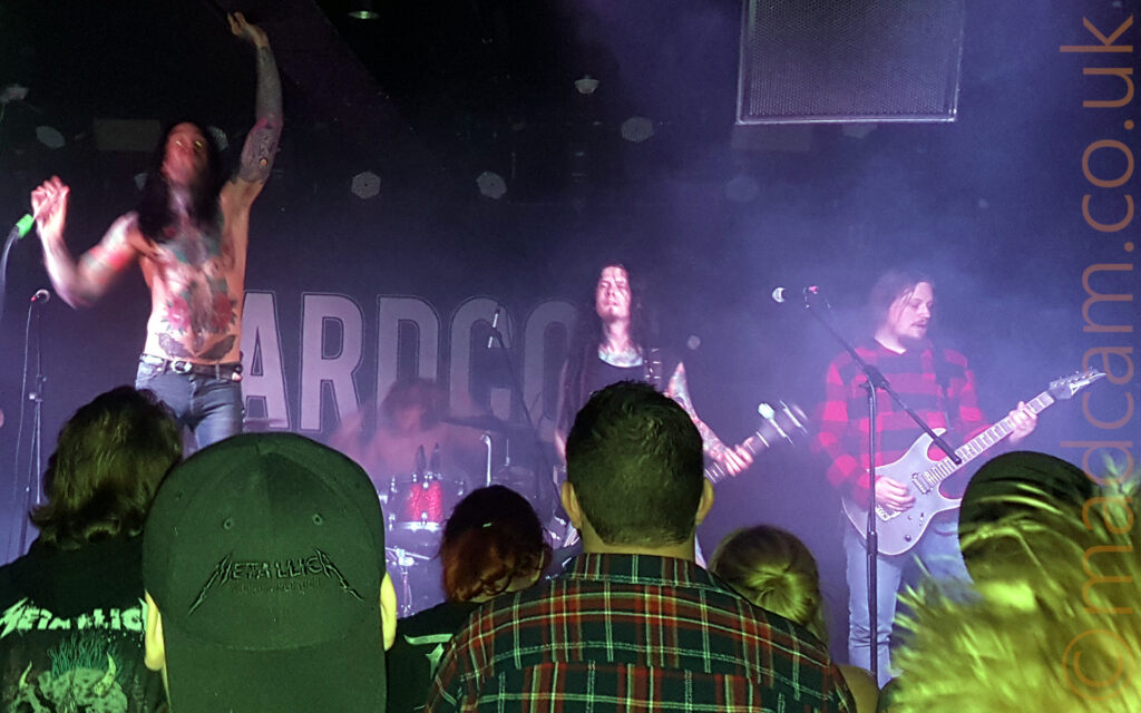 Five men performing on stage as a band. On the left is a man with long dark hair falling over his shoulders, wearing a black tank-top that expose his heavily tattooed arms, and a faded blue pair of jeans, playing an electric guitar while slightly hunched over. Further to the right is a man with long dark hair, his bare chest and arms covered in extravagant coloured tattoos, leaping into the air with his right hand holding a microphone, his left arm raised above his head. In the middle, right at the back, is a man sitting behind a red drum kit, pounding away on his instrument. Next is a man with long dark hair with a black tank top covering an extensively tattooed chest and leaving his tattooed arms exposed, playing an electric guitar. On the right is a man with his long dark hair tied back, wearing a red and black checked shirt, and playing a black electric guitar. The backs and heads of several people in the crowd can be seen in the foreground, partially obscuring the members of the band. Black walls and ceiling fill the rest of the frame, with the large white word "Hardcore" on the wall at the rear of the stage, and speakers hanging from the ceiling.