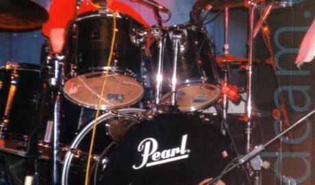 A man sitting behind a black drum kit, almost hidden by drums and cymbals, wearing a red t-shirt. There are white "Pearl" titles on the bass drum on the bottom of the kit. A black drape with an indistinct white logo hangs on the black wall on the back. A red light in the top right corner illuminates the chrome on the drum kit, as well as the drummer.