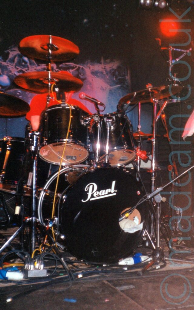 A man sitting behind a black drum kit, almost hidden by drums and cymbals, wearing a red t-shirt. There are white "Pearl" titles on the bass drum on the bottom of the kit. A black drape with an indistinct white logo hangs on the black wall on the back. A red light in the top right corner illuminates the chrome on the drum kit, as well as the drummer.