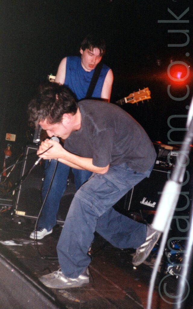 A man with short dark hair, wearing a grey t-shirt and dark blue trousers, hunched over, singing into a microphone he is holding in both hands. Another man, with short, floppy dark hair, wearing a sleeveless blue t-shirt and blue jeans, playing an electric guitar with his shoulders hunched over, is standing behind the singer. Amplifier cabinets sit on the floor of the stage in the background, with black walls and ceilings beyond them. A single red light stares balefully on the top right of the frame.