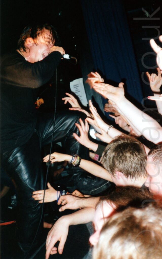 A man clad all in black, with straggly dark hair, screaming into a microphone in his right hand, as a wall of arms belonging to the crowd reach out towards him. Black walls fill the background.
