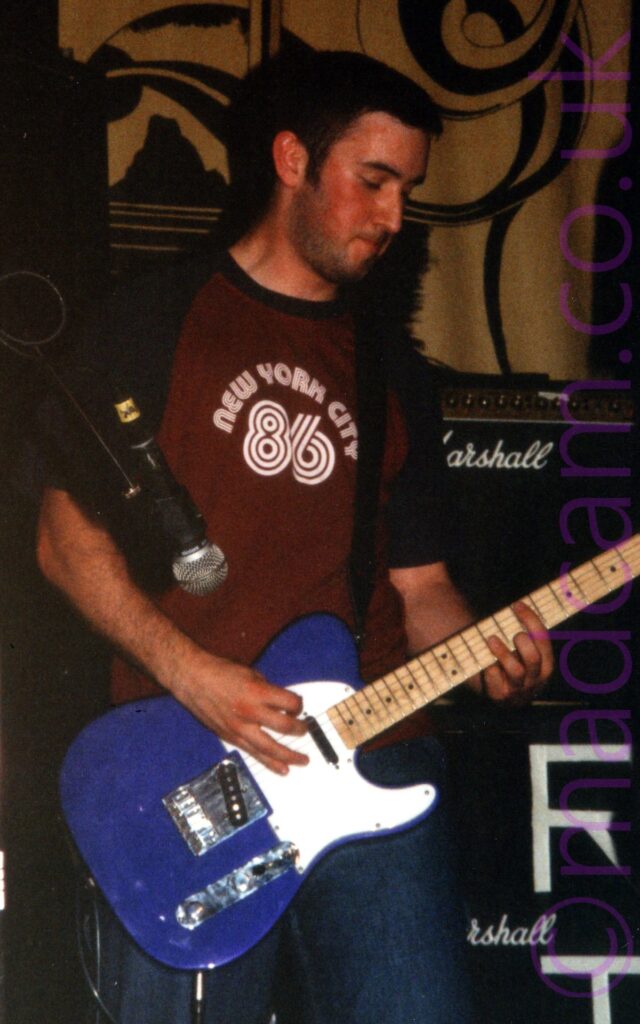 A man with short dark hair and short stubble on his face, wearing a maroon t-shirt with the words "New York City" and the number "86" emblazoned across the chest, playing a purple electric guitar. Stacks of black amplifiers with white "Marshall" text on the front fill the background at the bottom of the frame, with golden drapes covered in elaborate black patterns cover the walls at the top.