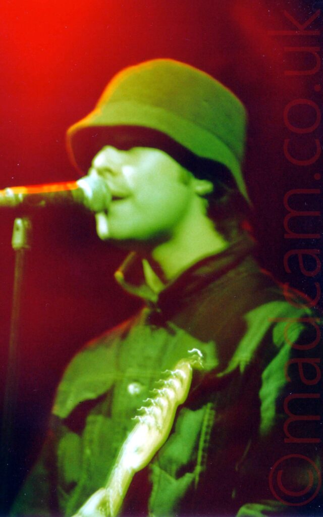 Head and shoulders shot of a man, wearing a dark coloured bucket hat and a denim jacket, playing an electric guitar while singing into a microphone on a stand. He is given a green cast by some stage lights, with blooms of red light filling the rest of the frame.