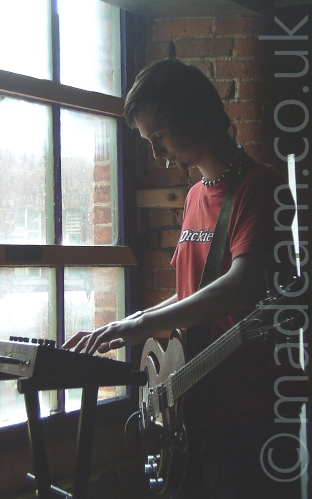 Almost moody shot of a man with short dark hair and goatee beard, wearing a red t-shirt with the white word "Dickies" across the front, with an electric guitar attached to a strap over his left shoulder hanging free in front of him. Instead of playing the guitar, he is playing a compact keyboard. A large window next to him on the left of the frame lets in plenty of light, but leaving everything not directly illuminated in shadow.