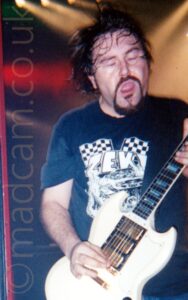 A man with mid-length dark hair, a dark goatee and moustache on his face, and wearing a dark blue t-shirt with a white print on the front. His mouth is open, and his eyes are closed, with his hair moving as he rocks out. He is playing a white electric guitar. White, yellow, and red shafts of light criss-cross the black background at the top of the frame, behind his head.