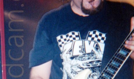 A man with mid-length dark hair, a dark goatee and moustache on his face, and wearing a dark blue t-shirt with a white print on the front. His mouth is open, and his eyes are closed, with his hair moving as he rocks out. He is playing a white electric guitar. White, yellow, and red shafts of light criss-cross the black background at the top of the frame, behind his head.