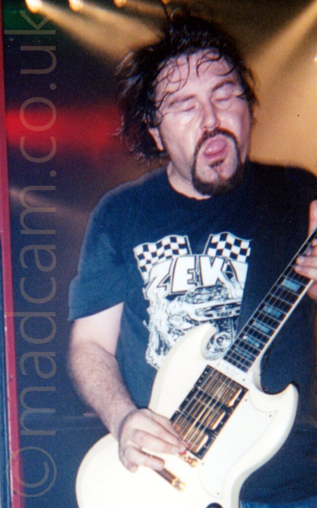 A man with mid-length dark hair, a dark goatee and moustache on his face, and wearing a dark blue t-shirt with a white print on the front. His mouth is open, and his eyes are closed, with his hair moving as he rocks out. He is playing a white electric guitar. White, yellow, and red shafts of light criss-cross the black background at the top of the frame, behind his head.
