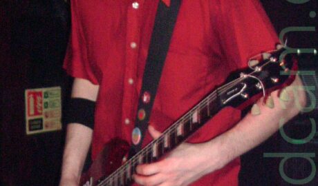 A man with lank, dark hair, and a little goatee beard, wearing a red shirt and a blue pair of jeans, playing a red electric guitar. There is a square, green sticker on his left leg, just below the pocket of his jeans. A couple of red signs on the wall on the left indicate where fire fighting equipment is stored, and a black and yellow strip near the top of the wall on the right.
