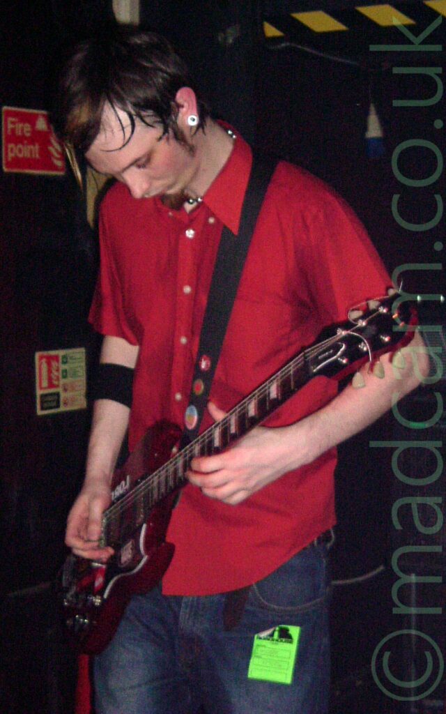A man with lank, dark hair, and a little goatee beard, wearing a red shirt and a blue pair of jeans, playing a red electric guitar. There is a square, green sticker on his left leg, just below the pocket of his jeans. A couple of red signs on the wall on the left indicate where fire fighting equipment is stored, and a black and yellow strip near the top of the wall on the right.
