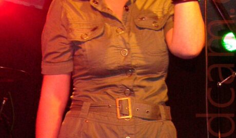 A woman with red hair cut in a bob, wearing a green shirt-style dress with press-studs up the front and tied with a green belt, holding a microphone on a stand in her left hand as she sings into it, while holding a megaphone low down in her right hand. A stagelight in the background on the left casts everything in red. A white suspended ceiling and black walls fill the rest of the frame.