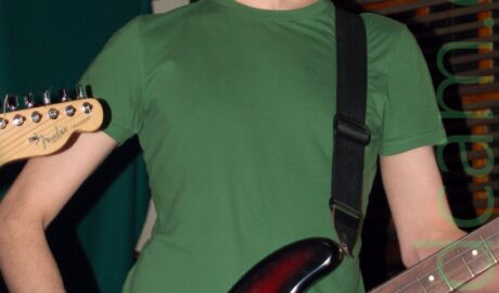 A man with shaggy brown hair, wearing a green t-shirt and blue jeans, blacking a black sunburst bass guitar with a white panel suspended from a black guitar strap over his left shoulder. He is looking quizzically at the camera. The head of an electric guitar is just coming into frame on the left. Brown wooden panels cover the wall behind him.