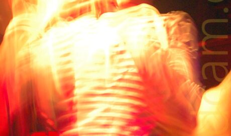 Long exposure photo of the singer with a band performing on a stage. He has a shaved head, and is wearing a black and white, thin banded long-sleeved t-shirt, and a blue pair of jeans. He is holding a microphone in his right hand, as he sings into it. A long exposure and his movement means that he is blurred and indistinct, with secondary images, including a rather ghostly face, all around him. Yellow stage lights illuminate the whole image, mixed with red, giving a rather spooky feel to the whole thing. Black walls and ceiling fill the background.
