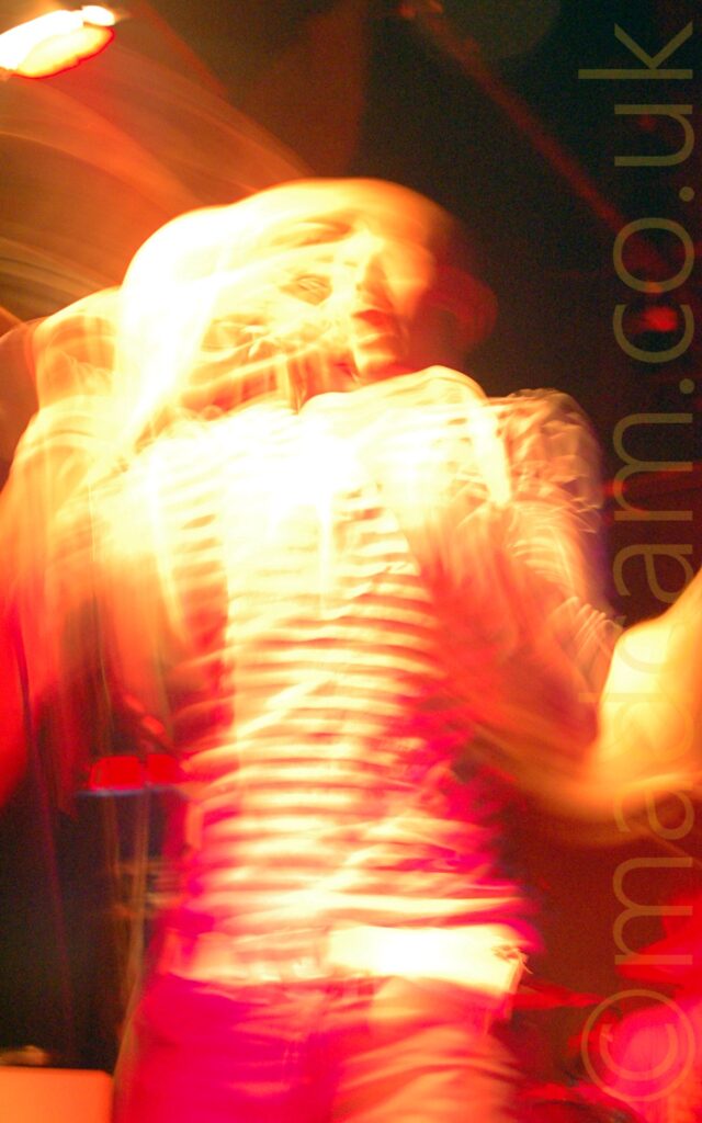 Long exposure photo of the singer with a band performing on a stage. He has a shaved head, and is wearing a black and white, thin banded long-sleeved t-shirt, and a blue pair of jeans. He is holding a microphone in his right hand, as he sings into it. A long exposure and his movement means that he is blurred and indistinct, with secondary images, including a rather ghostly face, all around him. Yellow stage lights illuminate the whole image, mixed with red, giving a rather spooky feel to the whole thing. Black walls and ceiling fill the background.