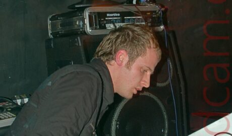A man with short dark hair, wearing a black rain jacket over a blue pair of jeans, hunched over slightly, playing a keyboard with both hands. The keyboard is sitting on a white wooden cabinet. a stack of amplifiers are stacked against the black wall in the background, with a red moving LED display at the top.