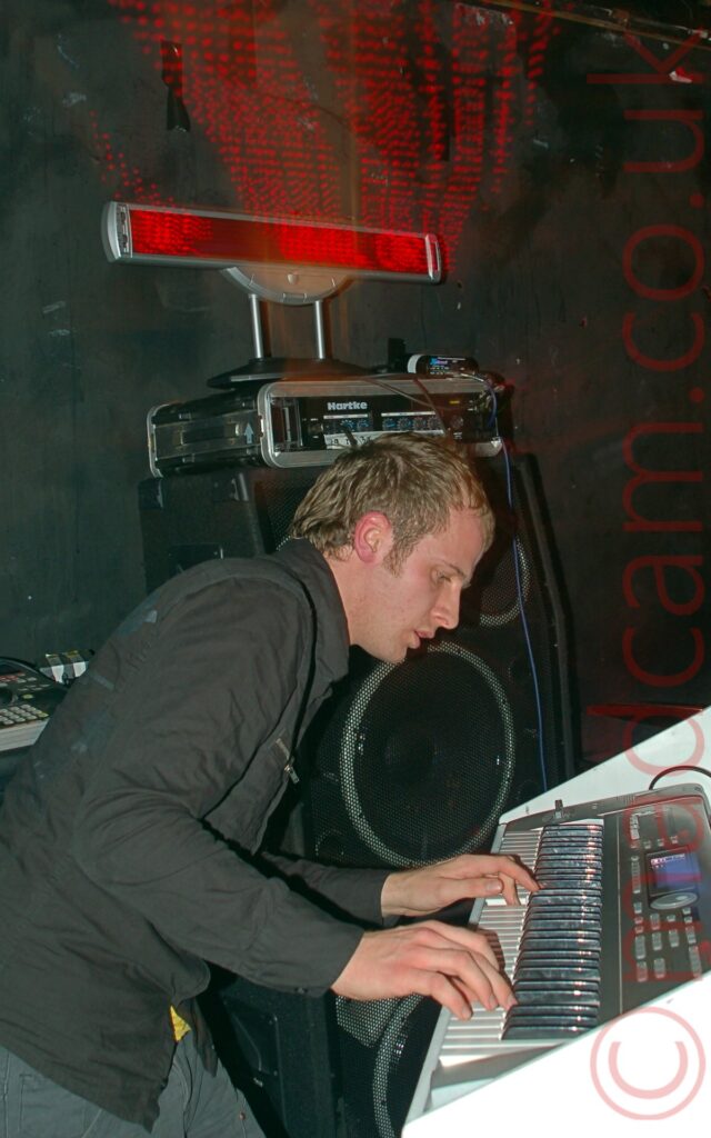 A man with short dark hair, wearing a black rain jacket over a blue pair of jeans, hunched over slightly, playing a keyboard with both hands. The keyboard is sitting on a white wooden cabinet. a stack of amplifiers are stacked against the black wall in the background, with a red moving LED display at the top.